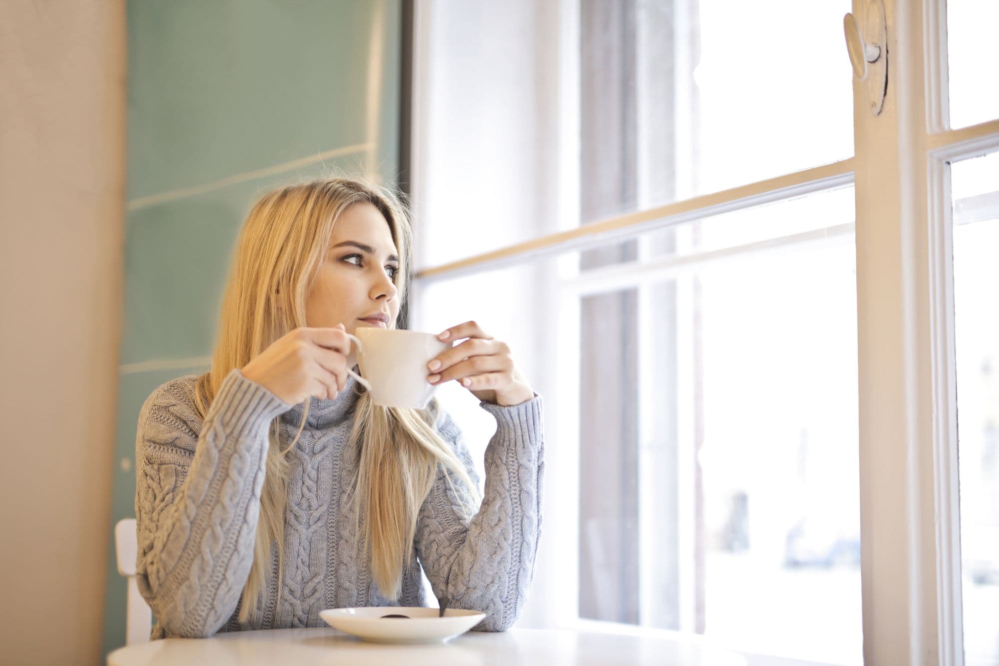 A woman holding a cup by a tall window, looking out